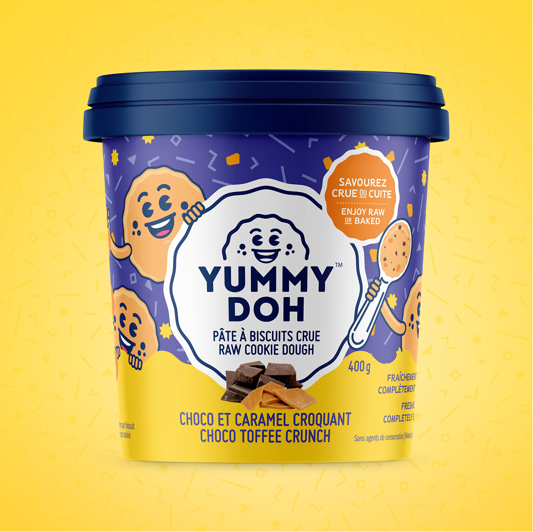 Un pot de 400 g de pâte à cookies crue « Yummy Doh », saveur « Choco & Caramel Croquant », avec un couvercle bleu et des mascottes en forme de cookies sur un fond violet et jaune. L'étiquette indique qu'elle peut être dégustée crue ou cuite au four.