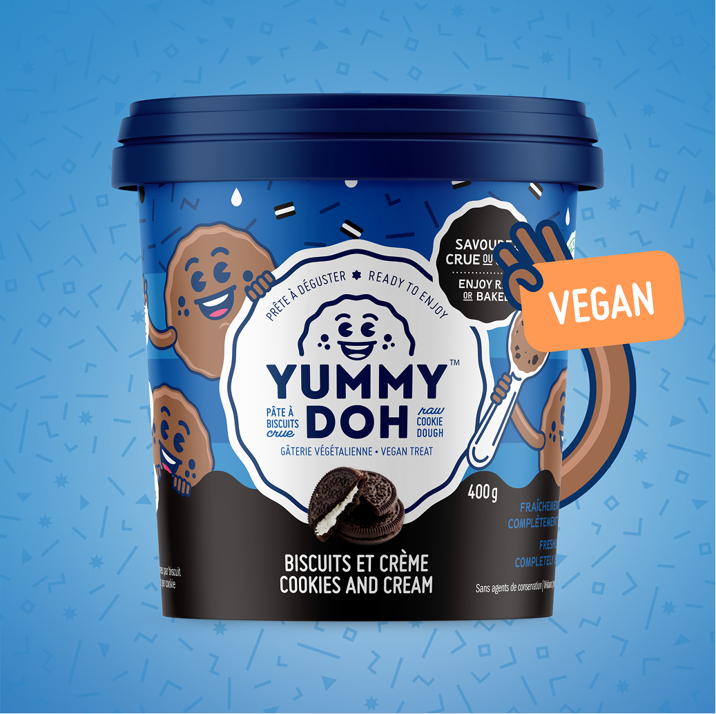 Un pot bleu de pâte à cookies végétalienne « Yummy Doh », au goût de biscuits et de crème, avec des cookies de dessin animé sur l'étiquette. Le couvercle est bleu foncé et une étiquette « VEGAN » y est apposée. Le fond est bleu clair et parsemé de paillettes.