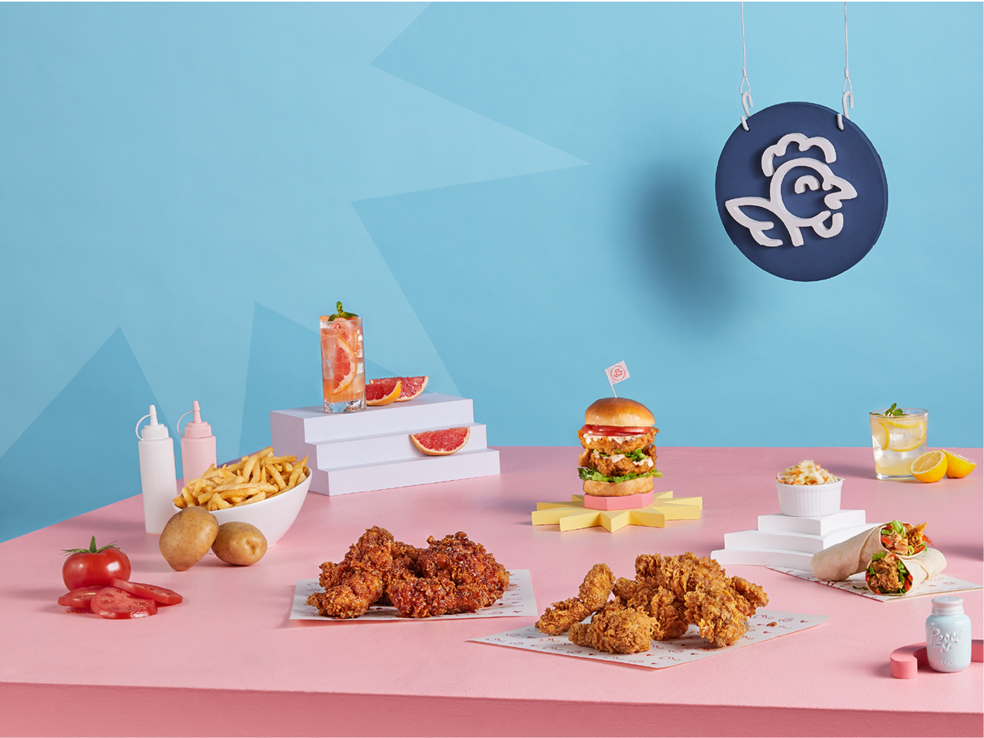 Une table chez Olivia’s Chicken est garnie de poulet frit, de frites, d’un hamburger, d’un wrap, de salade de chou, de sauces, de boissons, de tomates et de pommes de terre. Sur le fond bleu vif est accrochée une enseigne arborant le logo du restaurant spécialisé dans le poulet.