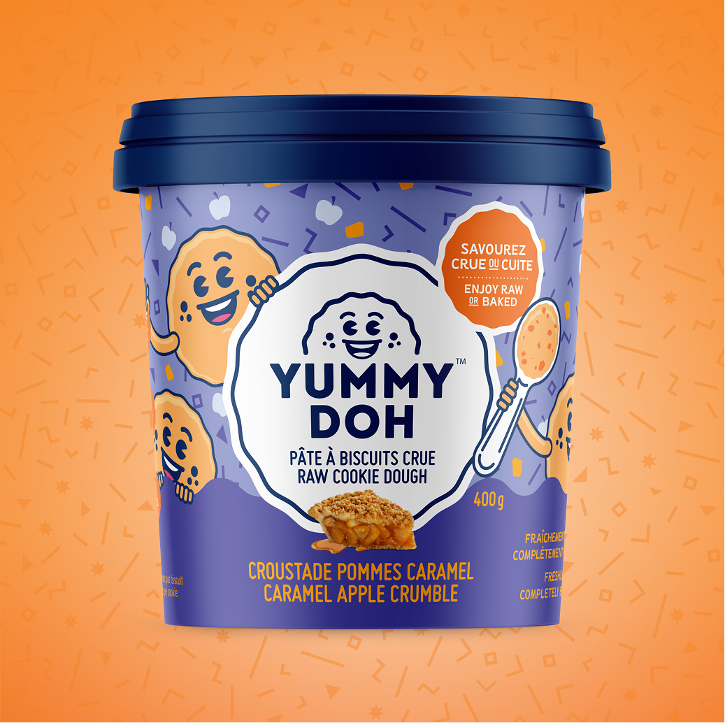 Un pot bleu et violet de pâte à cookies crue « Yummy Doh » au crumble de pomme au caramel, orné de mascottes souriantes en forme de cookies, sur un fond à motifs orange. L'étiquette indique que l'on peut la déguster crue ou cuite.