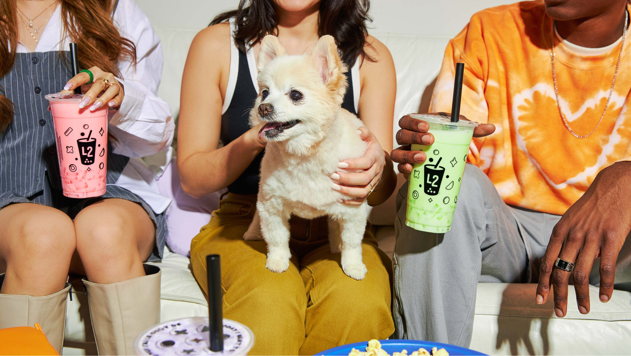 Trois personnes sont assises sur un canapé blanc, un bubble tea coloré de chez L2 Bubble Tea à la main, tandis qu'un petit chien au pelage duveteux est couché sur les genoux de la personne au milieu. Des en-cas sont disposés sur la table devant elles.