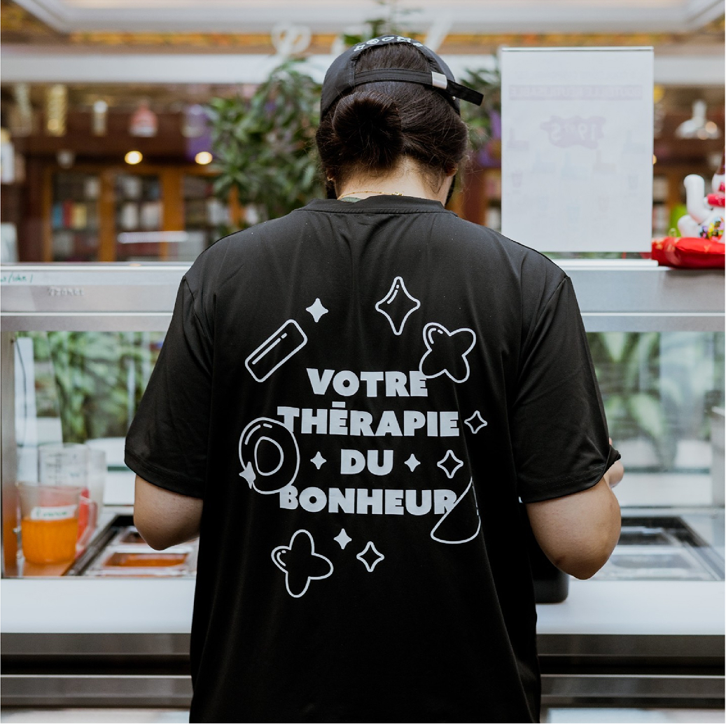 Une personne aux cheveux longs attachés en queue de cheval, vêtue d'un t-shirt noir sur lequel est inscrit en français « Votre thérapie du bonheur », se tient devant le comptoir d'un L2 Bubble Tea, dans un espace intérieur.