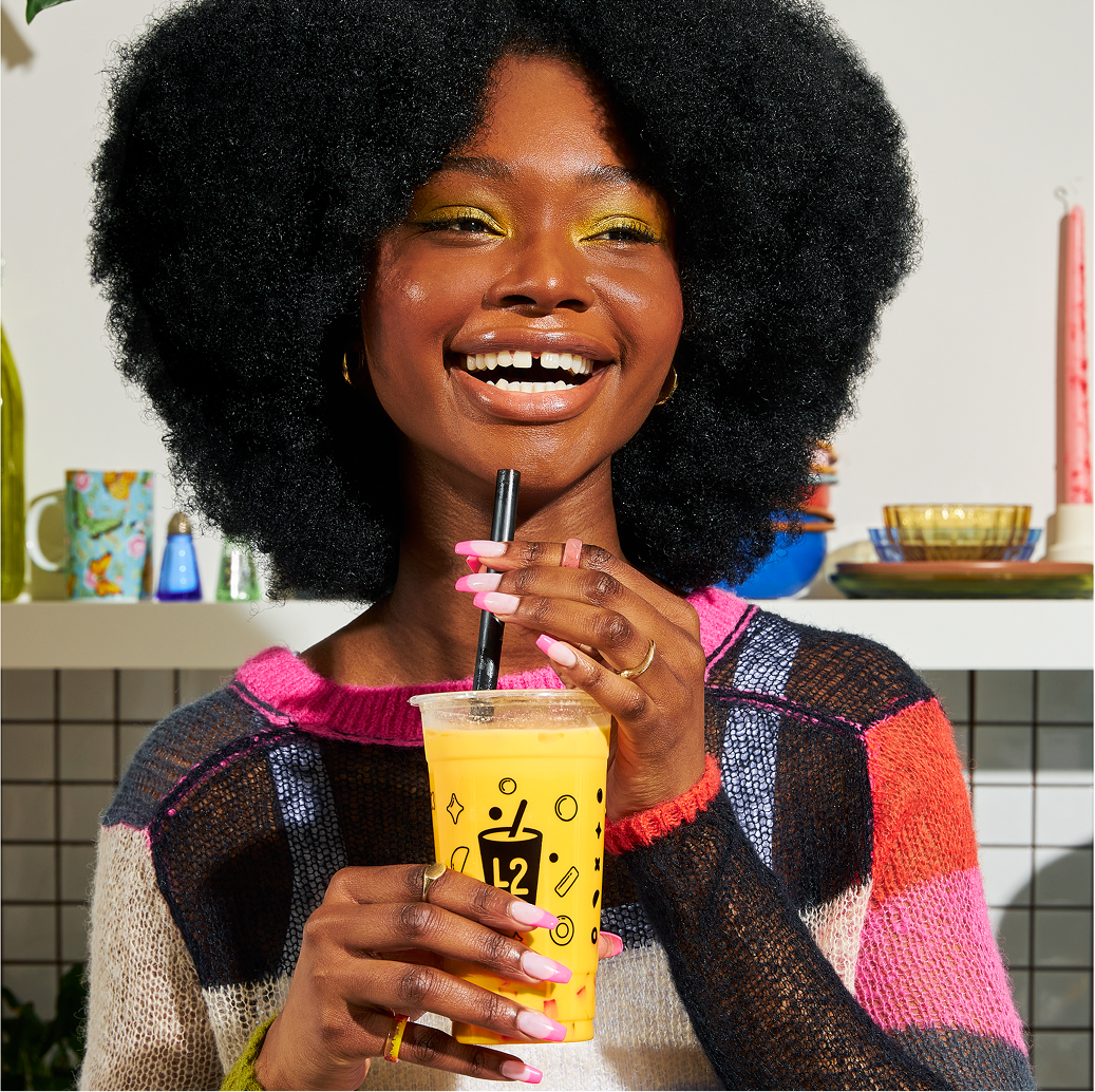 Une femme souriante, aux cheveux afro naturels, vêtue d'un pull coloré, tient un bubble tea jaune vif muni d'une paille noire. Elle se tient debout dans une cuisine animée, avec des assiettes et des tasses en arrière-plan.