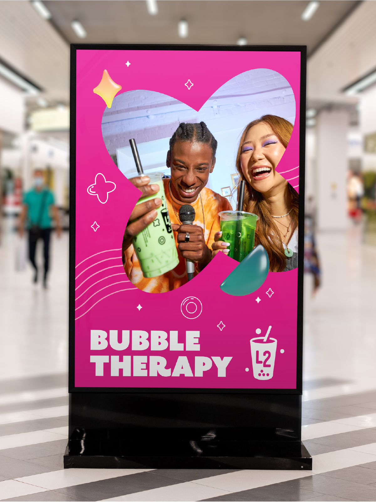 Un présentoir publicitaire dans un centre commercial affiche une affiche rose vif représentant deux personnes souriantes tenant des boissons au thé aux perles. Le texte indique « BUBBLE THERAPY », mettant en avant un partenariat créatif, avec une petite illustration de boisson et le logo « L2 » en bas.