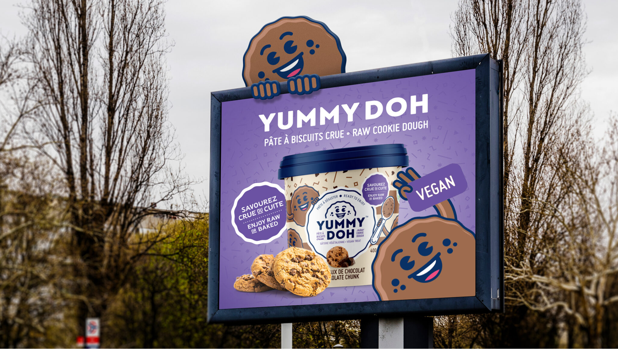 Une publicité sur panneau d'affichage pour la pâte à cookies végane « Yummy Doh », mettant en avant un pot du produit, des cookies illustrés et un personnage souriant en forme de cookie qui jette un coup d'œil par-dessus le bord. La publicité met en avant les mentions « végane » et « à déguster crue ou cuite ».