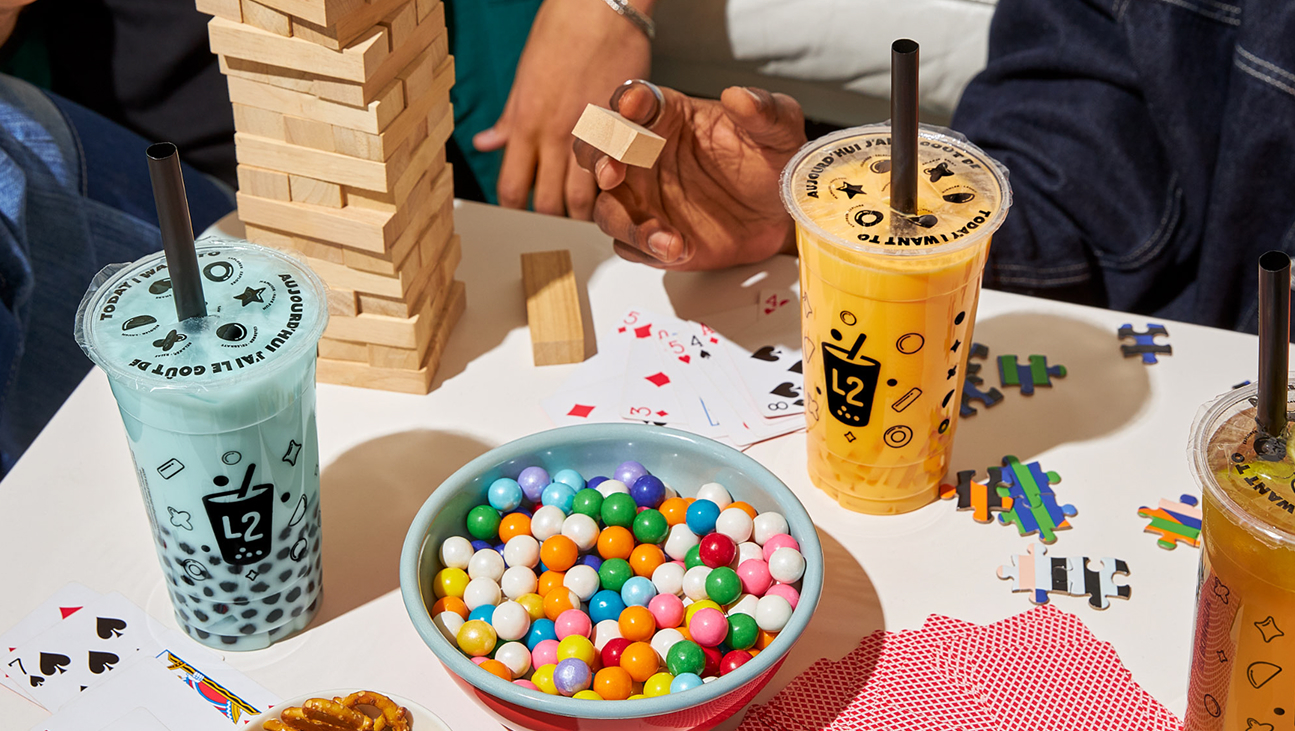 Un groupe de personnes joue au Jenga et à des jeux de cartes autour d'une table sur laquelle sont disposés des boissons au thé aux perles, un bol rempli de boules de chewing-gum colorées, des pièces de puzzle éparpillées et un jeu de cartes.