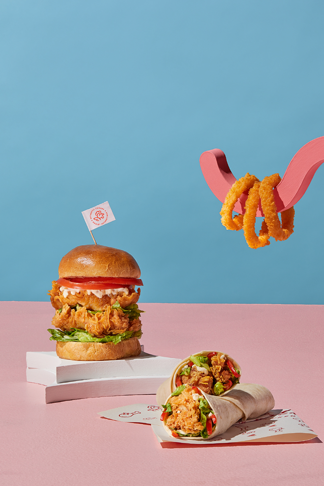 Un sandwich au poulet frit, garni de laitue et de tomate, trône sur des boîtes, à côté d'un wrap au poulet émincé de chez Olivia’s Chicken. Une fourchette rose retient trois rondelles d'oignon au-dessus d'un fond rose sur fond bleu, illustrant ainsi l'expérience authentique du poulet.