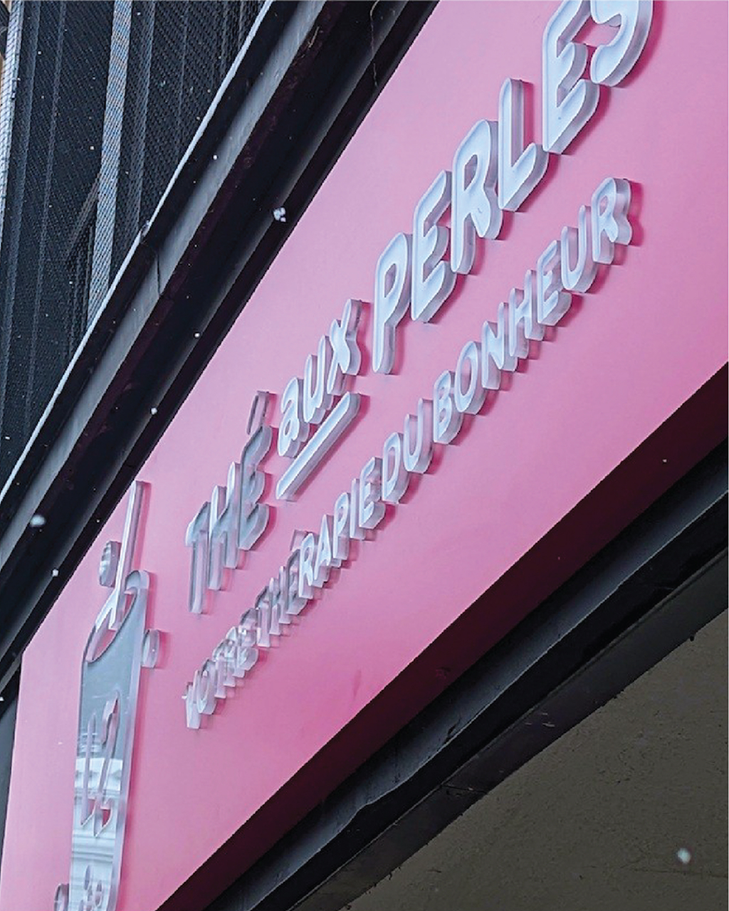 Une enseigne rose portant les inscriptions « THÉ aux PERLES » et « VOTRE THÉRAPIE DU BONHEUR » en lettres blanches en relief, vue en contre-plongée et sous un angle oblique. L'enseigne comporte l'illustration d'un verre de bubble tea, soulignant ainsi qu'il s'agit de l'adresse incontournable pour déguster du bubble tea.