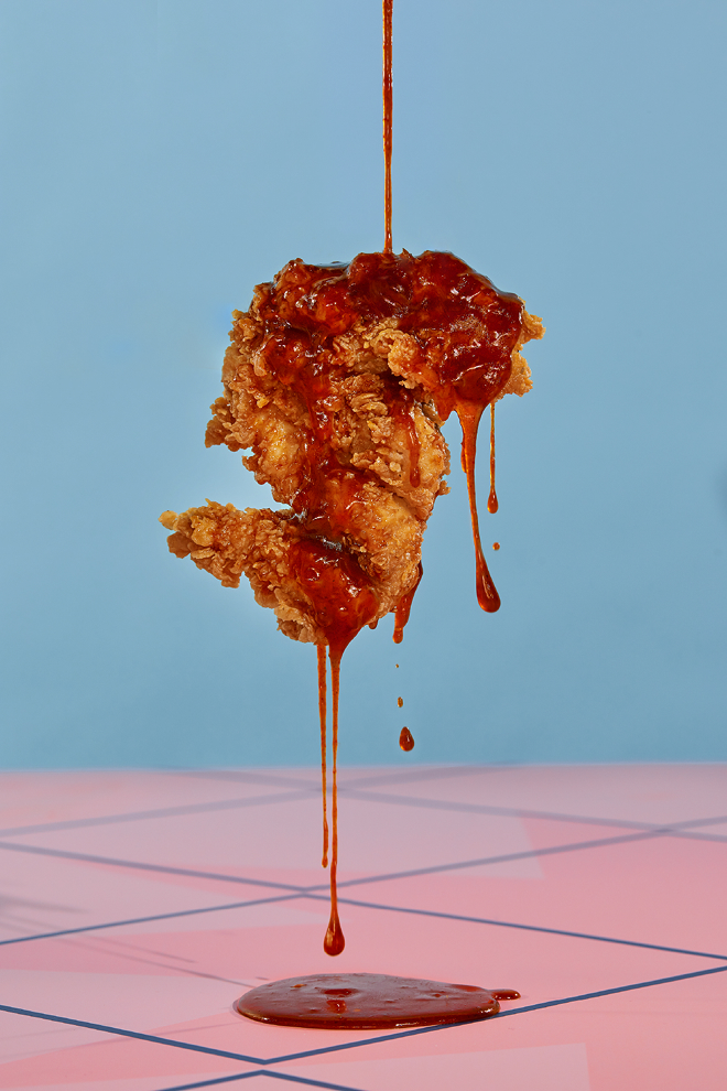Un pilon de poulet frit et croustillant de chez Olivia's Chicken est suspendu au-dessus d'une surface rose à motifs quadrillés, tandis qu'une sauce rouge épaisse s'en écoule, formant une petite flaque en dessous. L'arrière-plan est d'un bleu clair uni.