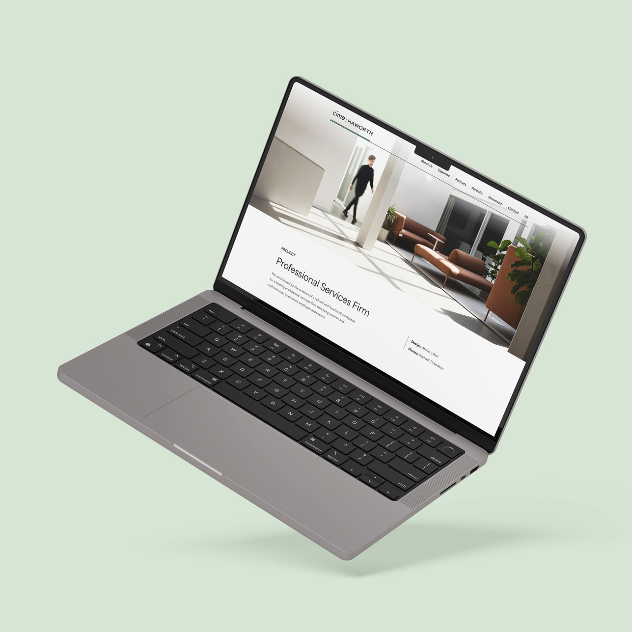 Un ordinateur portable gris se détache sur un fond vert clair, affichant un site web minimaliste avec un intérieur de bureau moderne, des plantes et une personne qui marche à l'arrière-plan.