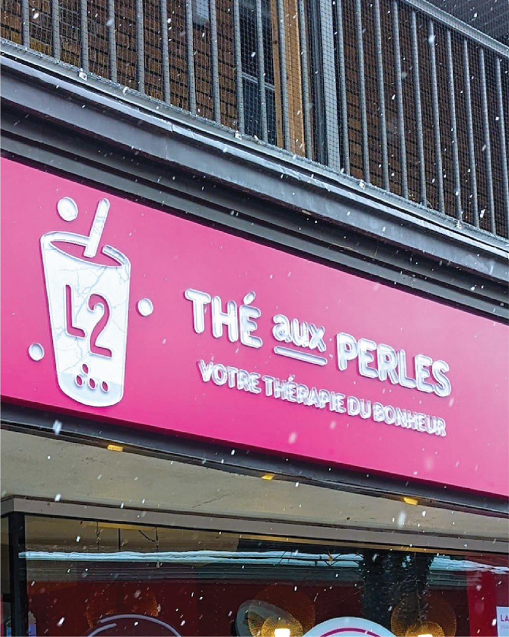 Une enseigne rose, ornée d'un logo représentant un verre de thé aux perles et portant les inscriptions « THÉ aux PERLES » et « VOTRE THÉRAPIE DU BONHEUR » en français, se détache sur fond de neige, vous invitant à profiter d'un moment de détente autour d'un thé aux perles.