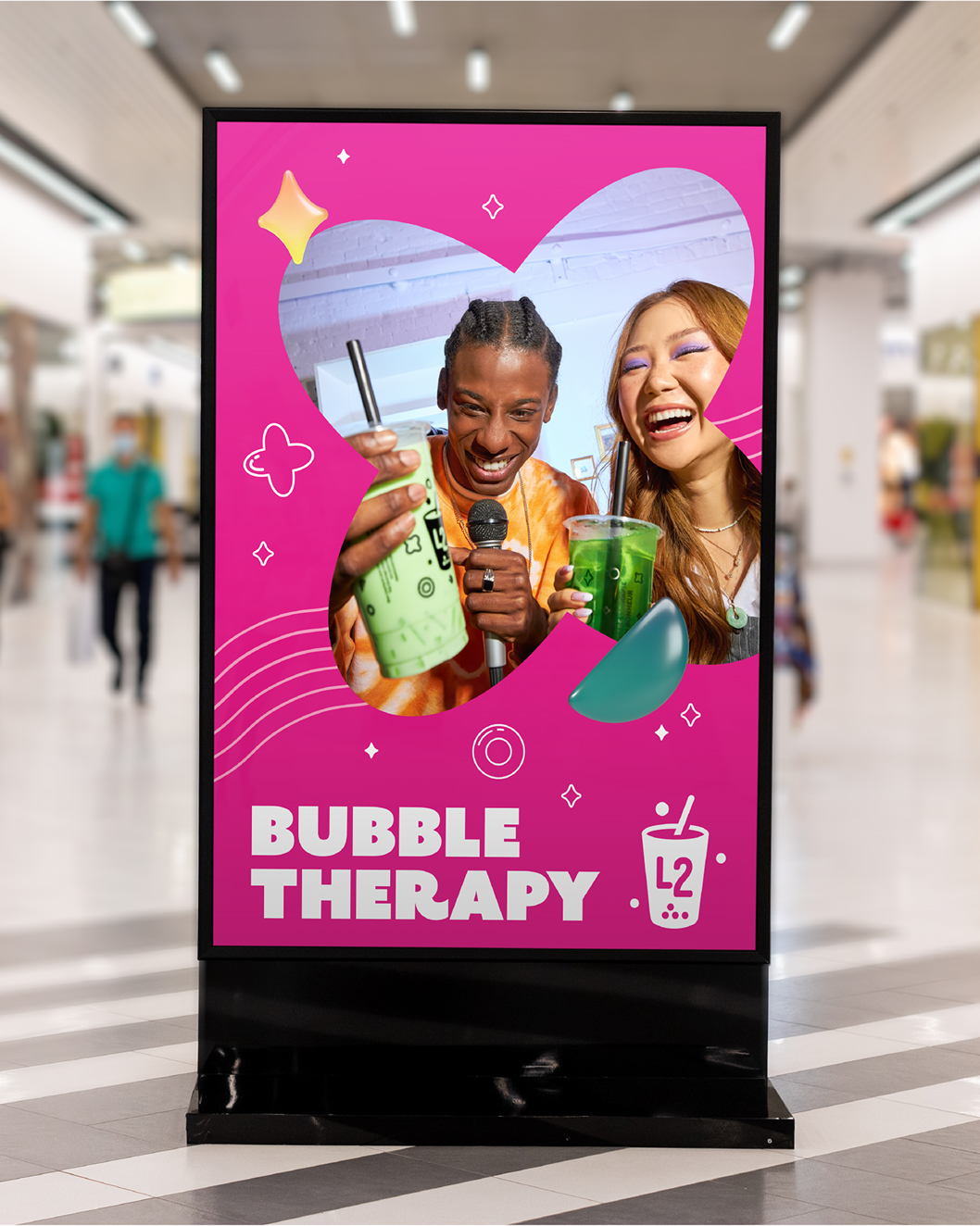 Une publicité rose dans un centre commercial met en scène deux personnes souriantes tenant chacune un bubble tea. Le texte indique « Bubble Therapy », accompagné d'une icône représentant un bubble tea et du logo L2. Des cœurs et des formes graphiques ludiques créent une ambiance amusante et accueillante pour les amateurs de bubble tea.