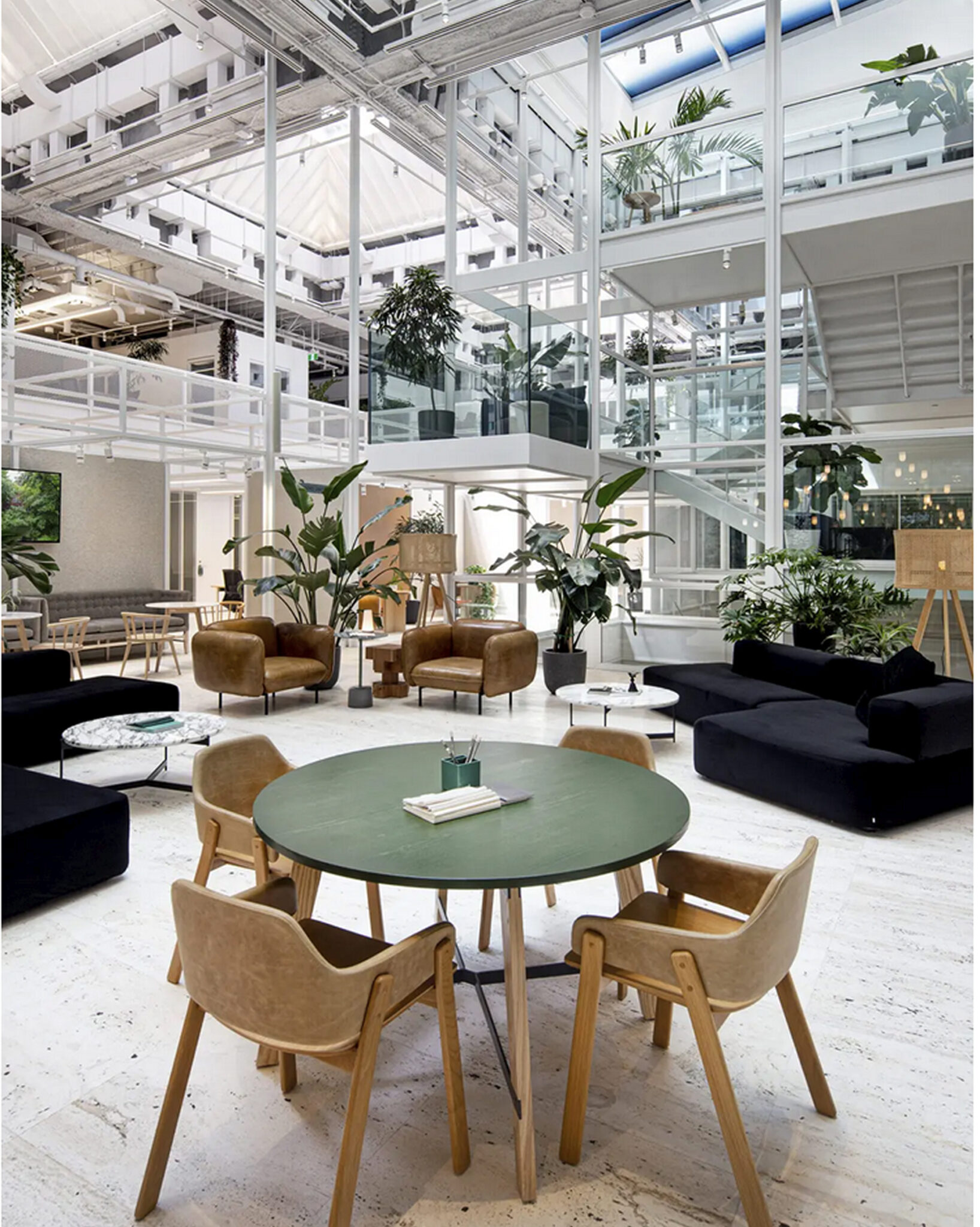 Un salon de bureau moderne et spacieux, doté de grandes baies vitrées, de plantes vertes, de canapés noirs, de fauteuils marron, d'une table ronde verte et d'un escalier blanc menant à un étage supérieur baigné de lumière naturelle.