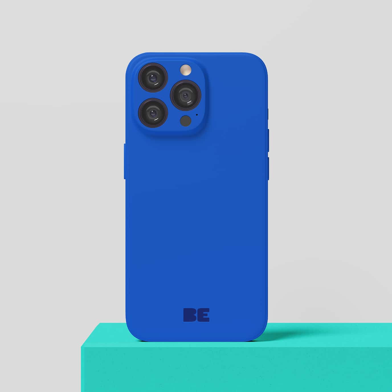 Un smartphone doté d'une coque bleu vif est posé à la verticale sur un socle turquoise, devant un fond gris clair uni. Le mot « BE » est imprimé au bas de la coque.