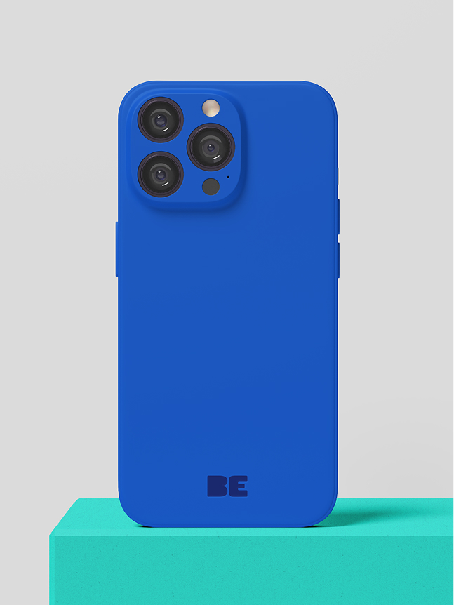 Un smartphone bleu, équipé de trois caméras à l'arrière, est posé à la verticale sur un bloc turquoise, devant un fond gris clair uni. L'inscription « BE » est imprimée au bas du dos de l'appareil.