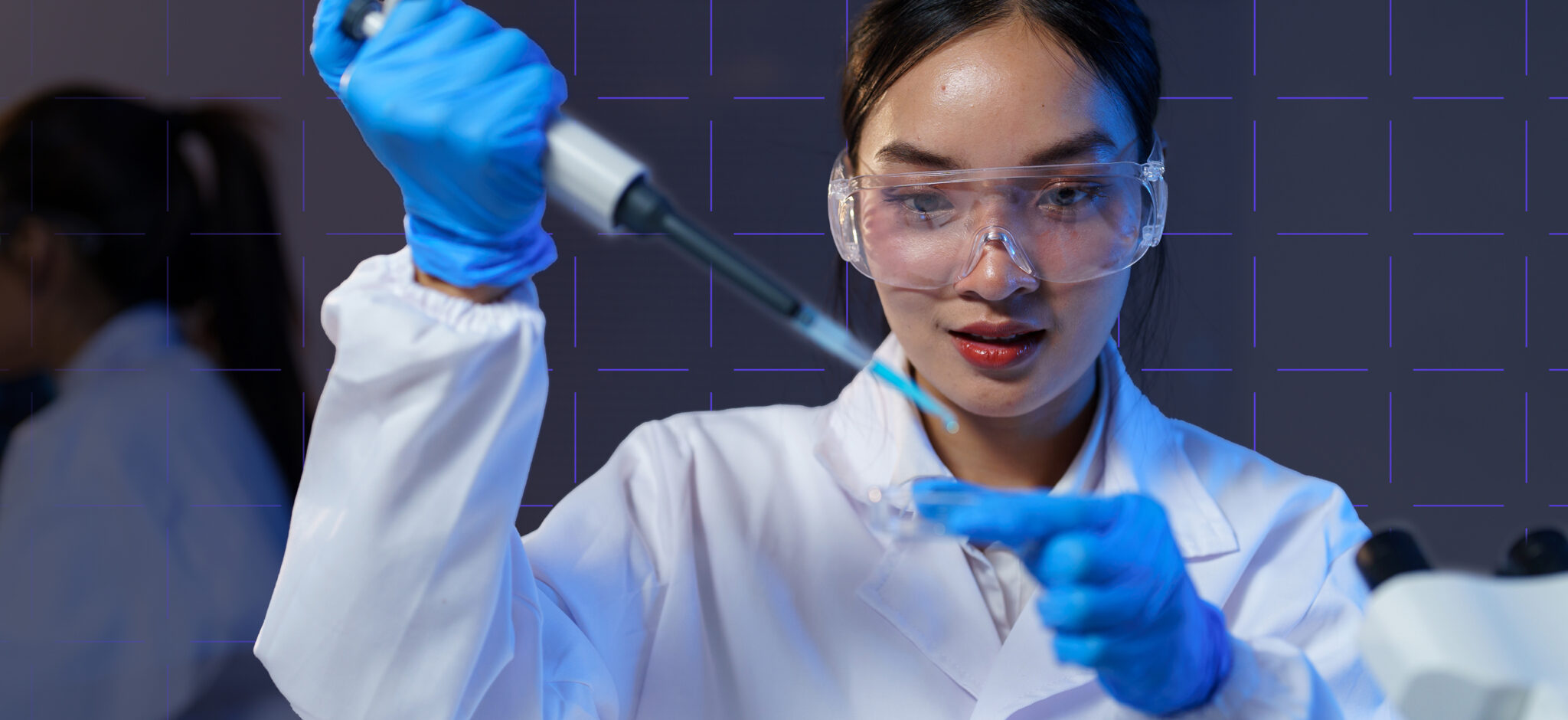 Dans un laboratoire, un scientifique portant des lunettes de protection et des gants bleus utilise une pipette pour transférer un liquide dans un tube à essai. On aperçoit une autre personne qui travaille à l'arrière-plan.