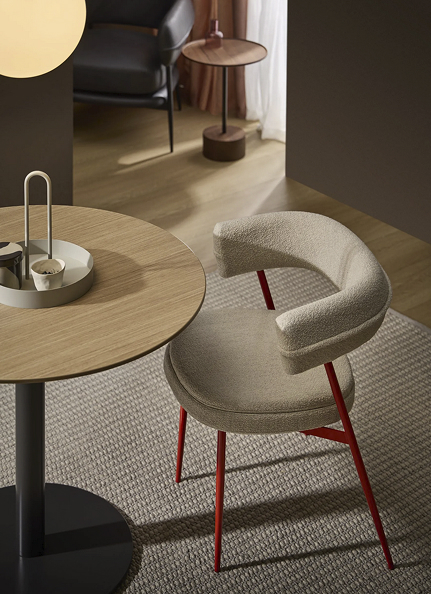 Une salle à manger moderne avec une table ronde en bois, une chaise rembourrée beige aux pieds métalliques rouges, un tapis texturé et une petite table d'appoint à l'arrière-plan. Un éclairage chaleureux crée une ambiance douillette.