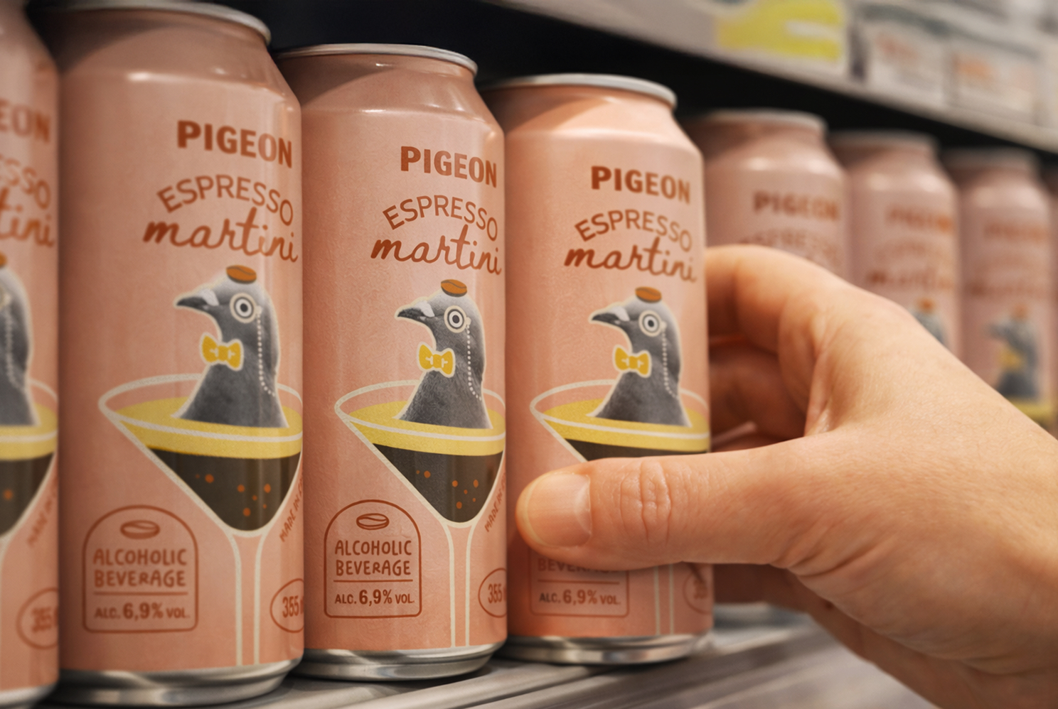 Une main s'empare d'une canette de « Pigeon Espresso Martini », dont l'étiquette rose représente un pigeon dans un verre à martini. D'autres canettes arborant le même motif sont alignées sur le rayon du magasin.