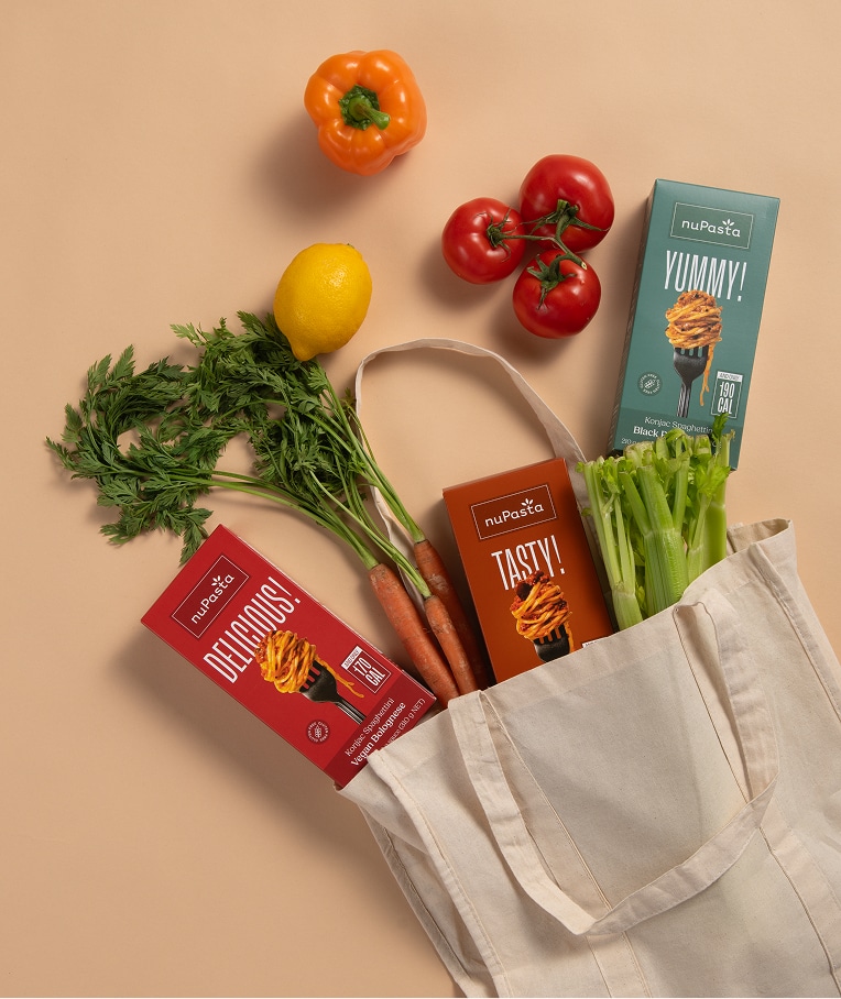 Un sac en tissu réutilisable contenant du céleri, des carottes, un citron et trois boîtes de nuPasta est posé sur une surface beige. À côté se trouvent un poivron jaune et une grappe de tomates avec leurs tiges.