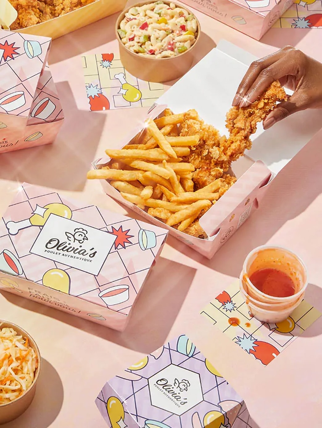 Une main trempe un morceau de poulet frit dans la sauce posée à côté d'une barquette de frites. D'autres barquettes, des saladiers de salade de pâtes et de salade de chou sont disposés sur la table rose, tous arborant le logo coloré d'Olivia's.
