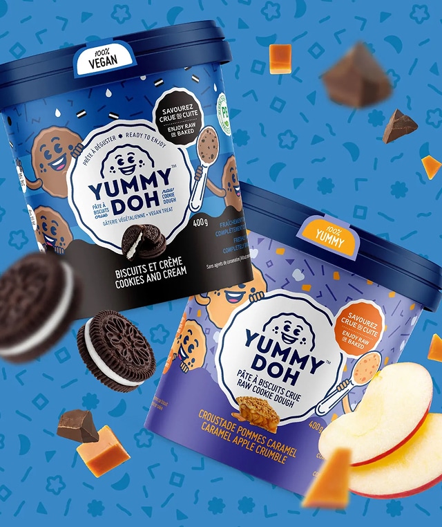 On voit deux pots de pâte à biscuits comestible « Yummy Doh », l'un au goût « Cookies and Cream » et l'autre au goût « Caramel Apple Crumble », sur fond illustré, avec des morceaux de biscuits, de caramel, de pomme et de chocolat en suspension.