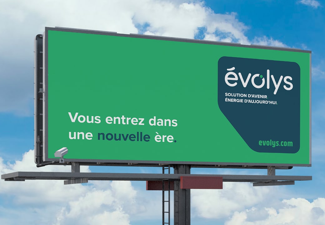 Un grand panneau publicitaire vert porte l'inscription suivante : « évolys. Solution d’avenir. Énergie d’aujourd’hui. Vous entrez dans une nouvelle ère. » L'adresse du site web, evolys.com, apparaît en bas. Le panneau se détache sur un ciel bleu parsemé de nuages.