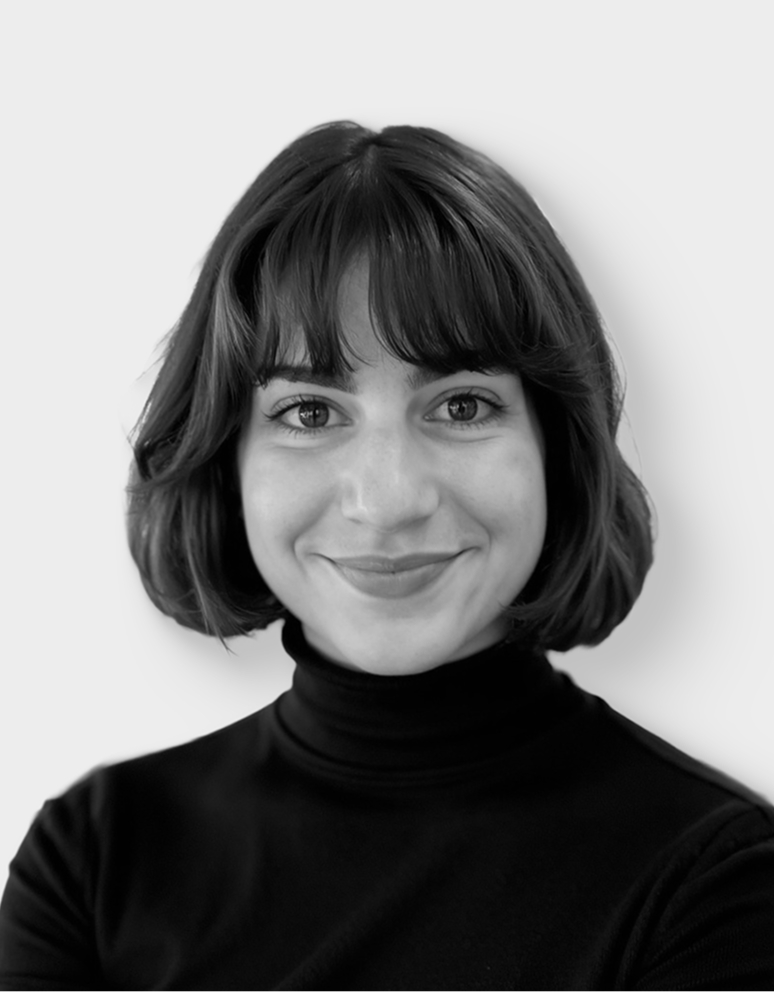 Une femme souriante, aux cheveux courts et foncés avec une frange, vêtue d'un pull à col roulé noir, pose devant un fond clair et uni.