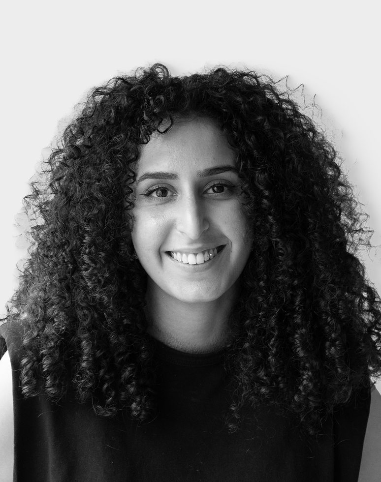 Une personne aux longs cheveux bouclés et foncés sourit à la caméra. Elle porte un haut sans manches de couleur foncée et pose devant un fond clair et uni.
