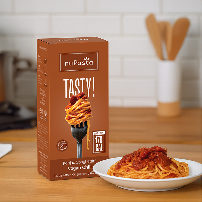 Une boîte de « nuPasta Konjac Spaghetti Vegan Chili » est posée sur un plan de travail en bois, à côté d'une assiette de spaghettis nappés de sauce tomate. Sur la boîte, les mentions « TASTY ! » et « 170 CAL » apparaissent en caractères gras.