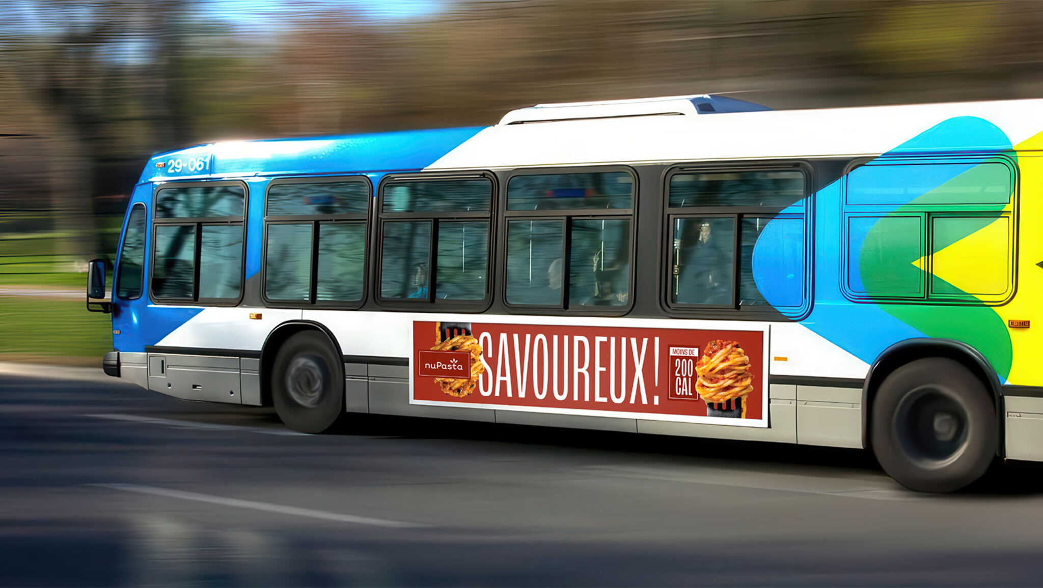 Un bus urbain en circulation arbore une publicité mettant en avant une pâtisserie sur laquelle figure en grosses lettres le mot « SAVOUREUX ! », dans le but de promouvoir un produit alimentaire. Le bus est peint dans des tons de bleu, blanc et vert.