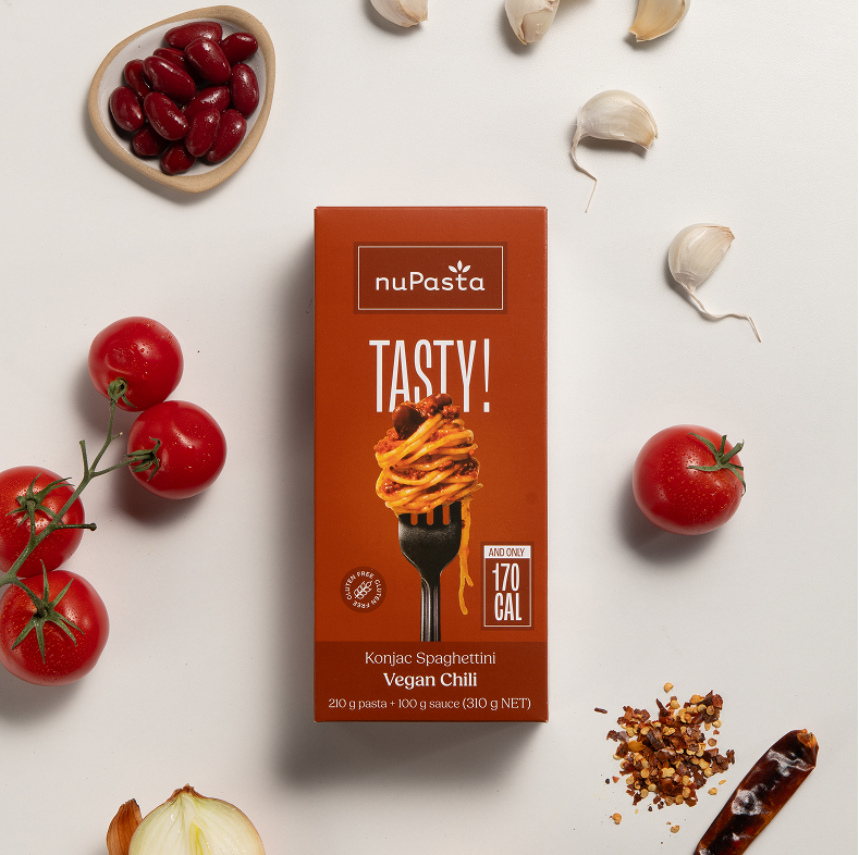 La boîte de « nuPasta Konjac Spaghetti Vegan Chili » est entourée de tomates fraîches, de haricots rouges, de gousses d'ail, d'oignon, de flocons de piment rouge et d'un brin de persil, le tout disposé sur un fond clair.