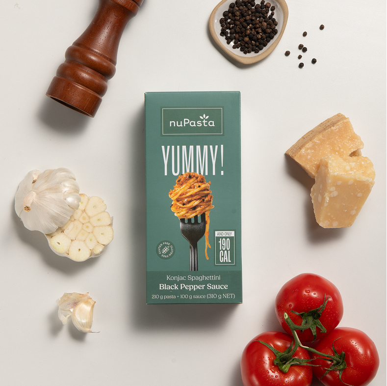 Une boîte de « nuPasta Konjac Spaghetti Black Pepper Sauce » est entourée d'une tête d'ail, de gousses d'ail en vrac, d'un moulin à poivre, de grains de poivre noir, de parmesan et de trois tomates, le tout posé sur une surface blanche.