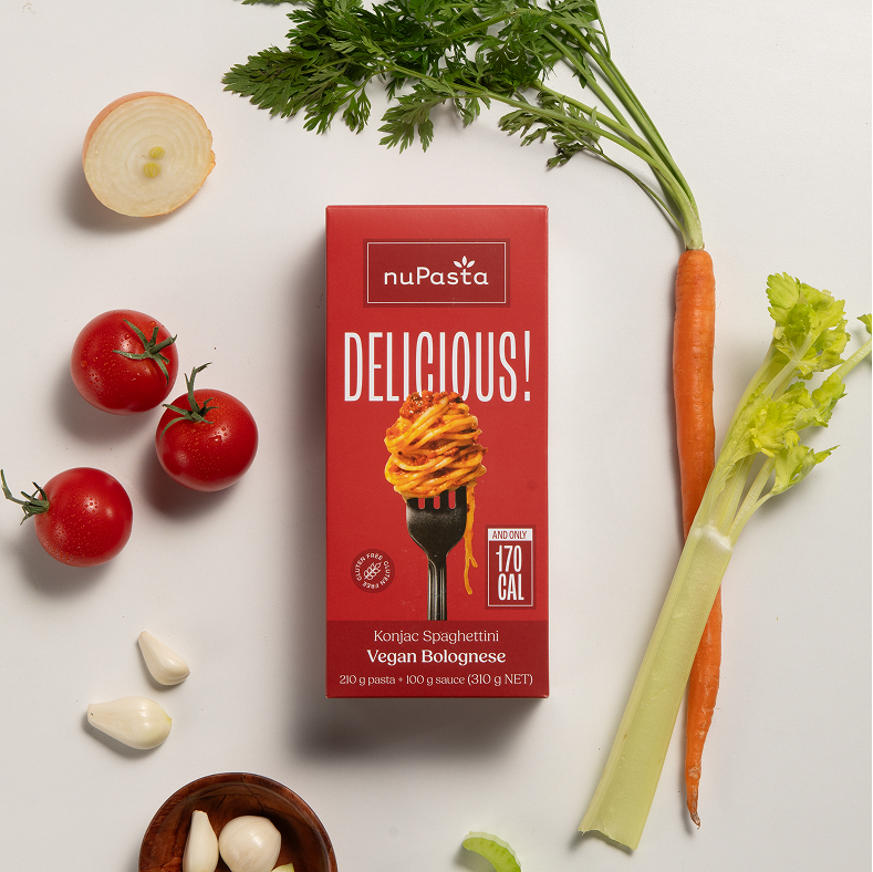 Une boîte de « nuPasta Konjac Spaghetti Vegan Bolognese » est entourée de légumes frais : une carotte, une branche de céleri, une grappe de tomates, un demi-oignon et des gousses d'ail, le tout disposé sur un fond blanc.