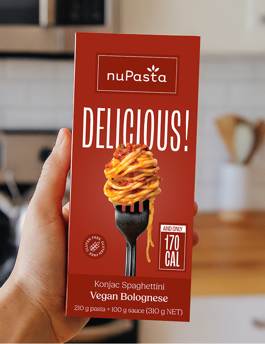 Dans une cuisine, une main tient une boîte rouge de « nuPasta Konjac Spaghettini Vegan Bolognese ». La boîte représente des spaghettis sur une fourchette, met en avant les mentions « Délicieux ! » et « 170 CAL », et précise la composition des pâtes et de la sauce.
