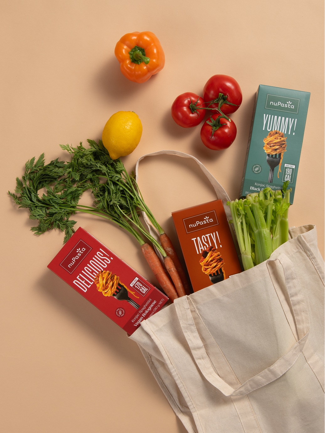 Un sac fourre-tout réutilisable rempli de carottes, de céleri et de trois boîtes de nuPasta. À côté du sac se trouvent un poivron jaune, un citron et une grappe de tomates, le tout sur fond beige.