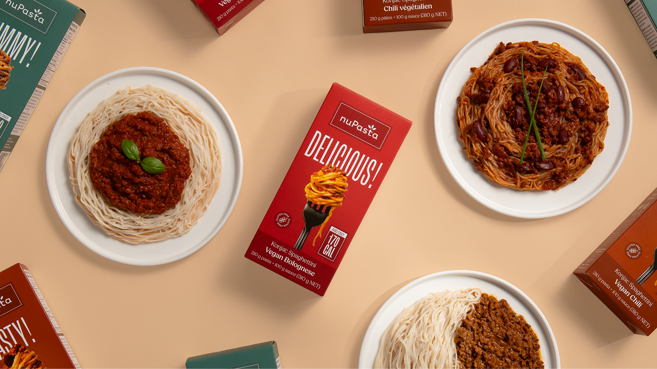Vue aérienne d'assiettes de spaghettis accompagnées de différentes sauces et de boîtes de « NUPASTA Vegan Bolognese » disposées parmi les assiettes, le tout sur un fond beige. Les plats sont agrémentés d'herbes fraîches.