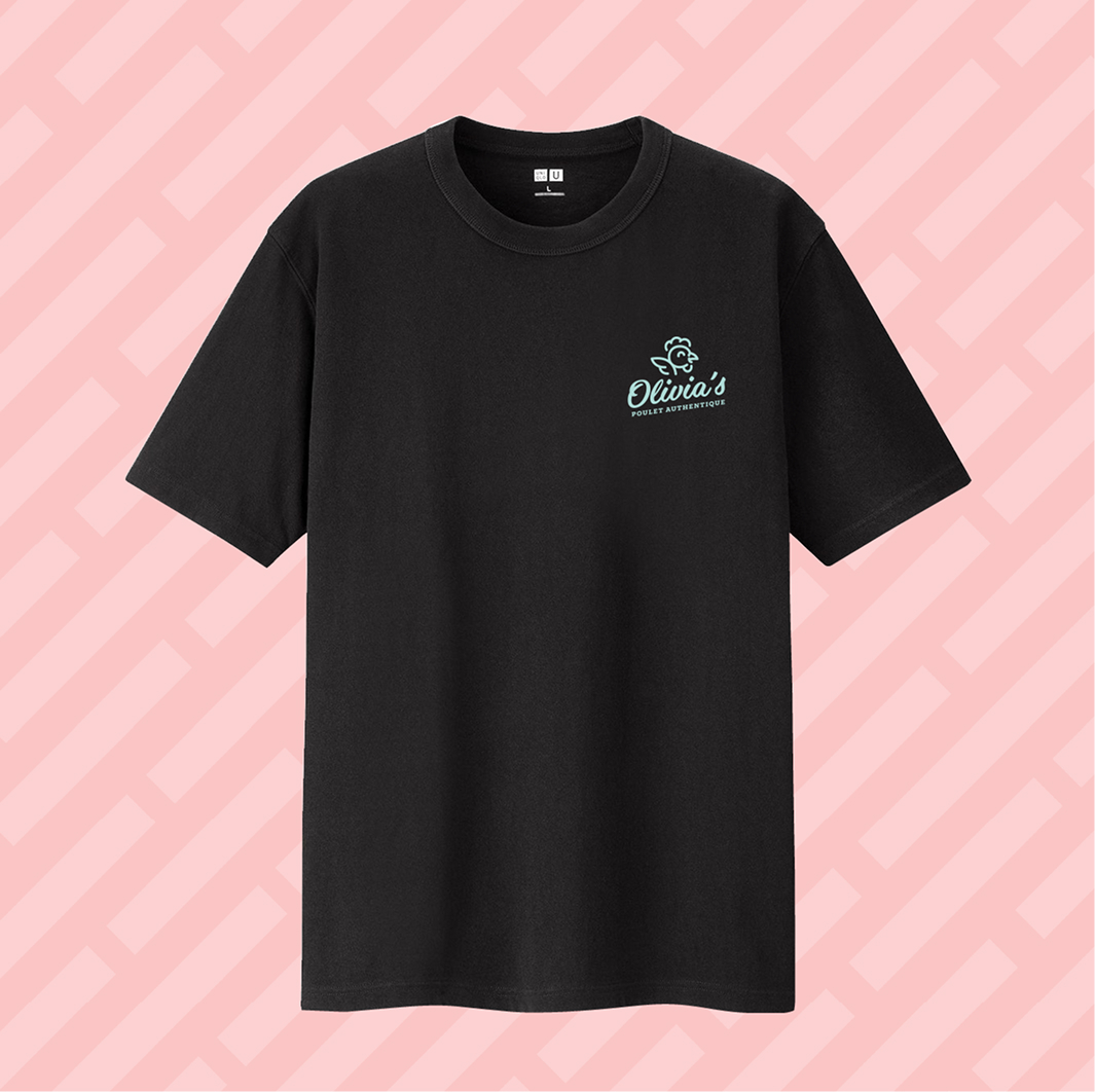 Un t-shirt noir à manches courtes orné d'un petit logo blanc et vert « Olivia’s Bakery & Cafe » représentant le poulet d'Olivia sur le côté gauche de la poitrine, sur fond rose à motifs géométriques.