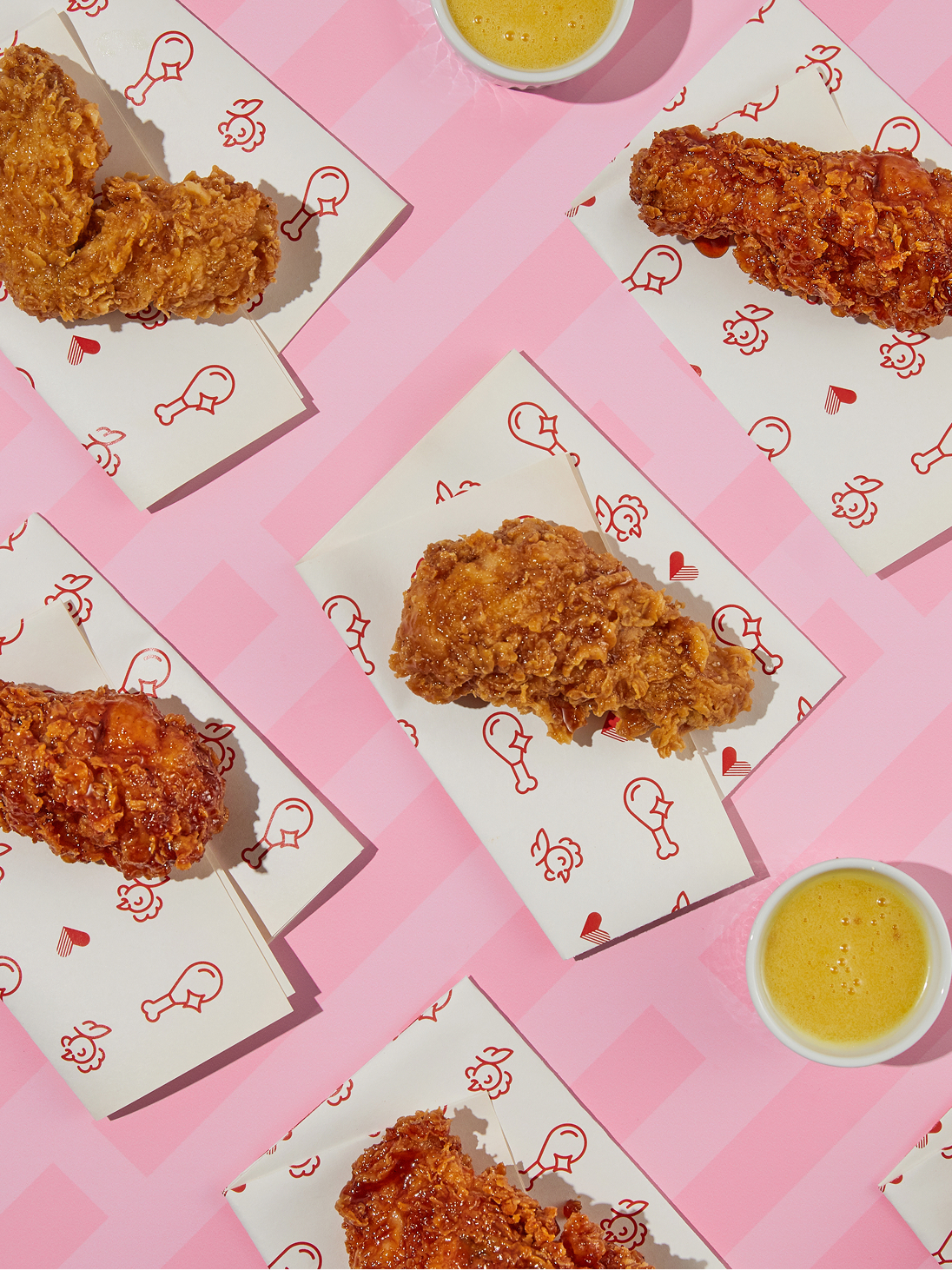 Des morceaux de poulet croustillants « Olivia’s » posés sur du papier à motifs, accompagnés de petites coupelles de sauce jaune, le tout disposé sur un fond à carreaux roses.