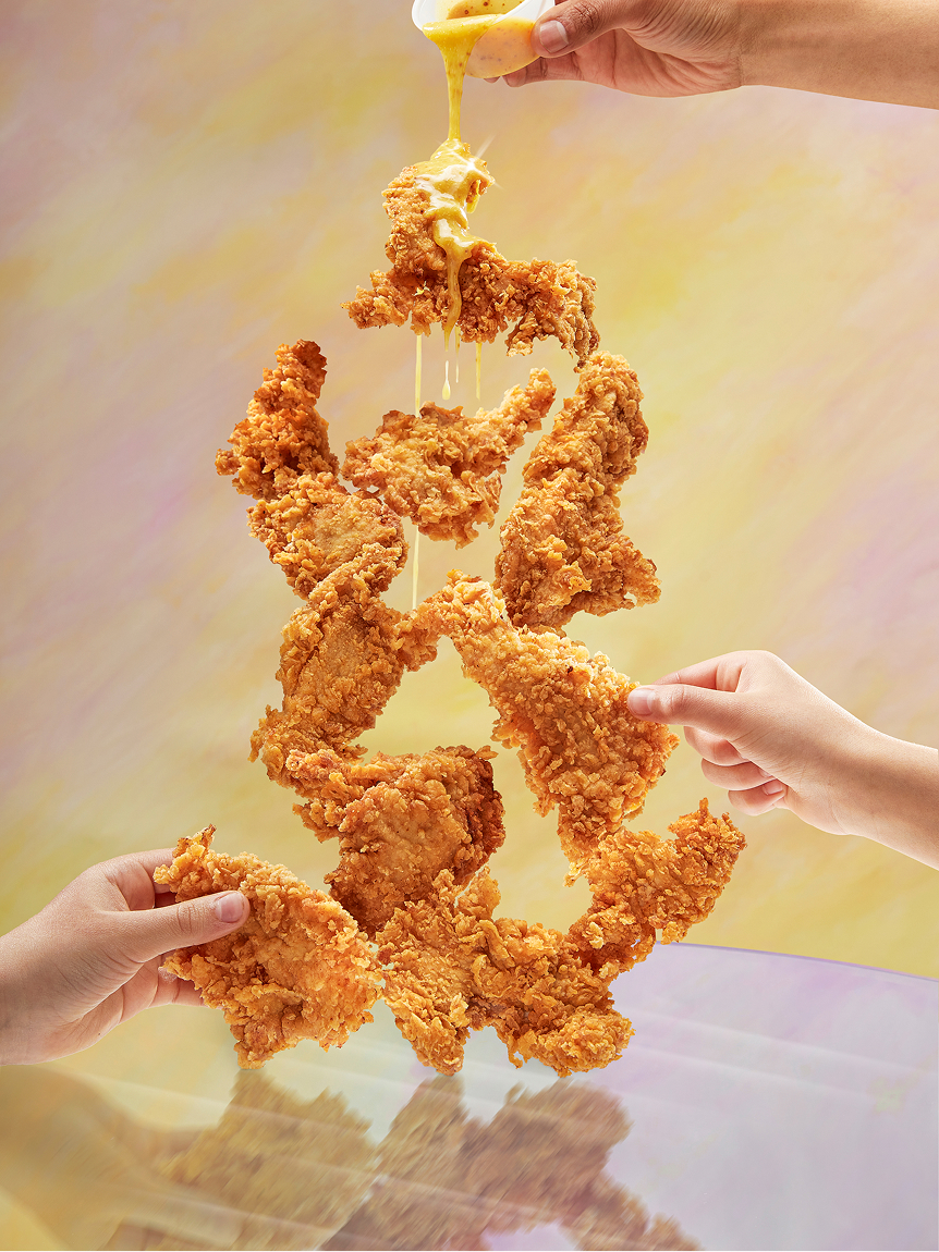 Trois mains tiennent des filets de poulet frits croustillants empilés à la verticale, tandis qu’une quatrième main verse de la sauce au fromage fondu par-dessus : une scène alléchante mettant en valeur le poulet d’Olivia, sur fond de couleurs pastel.