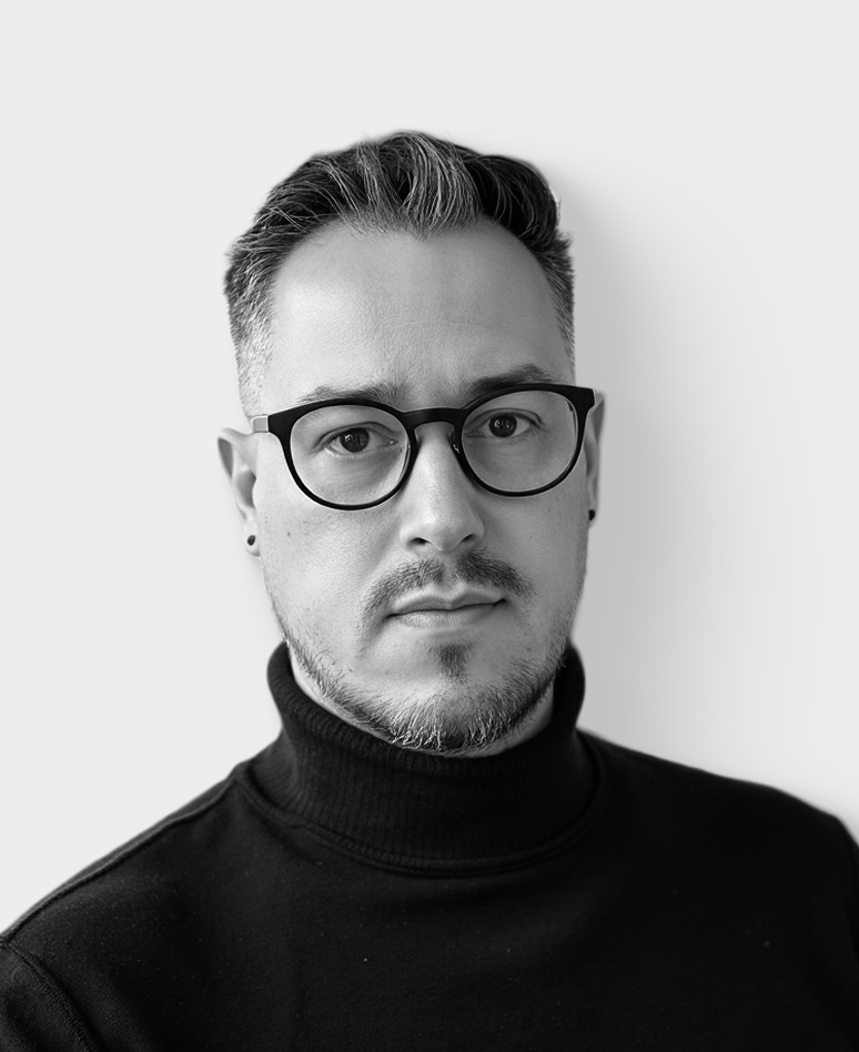 Un homme aux cheveux courts et foncés, à la barbe soignée et portant des lunettes, vêtu d'un pull à col roulé noir, regarde directement la caméra devant un fond clair et uni.
