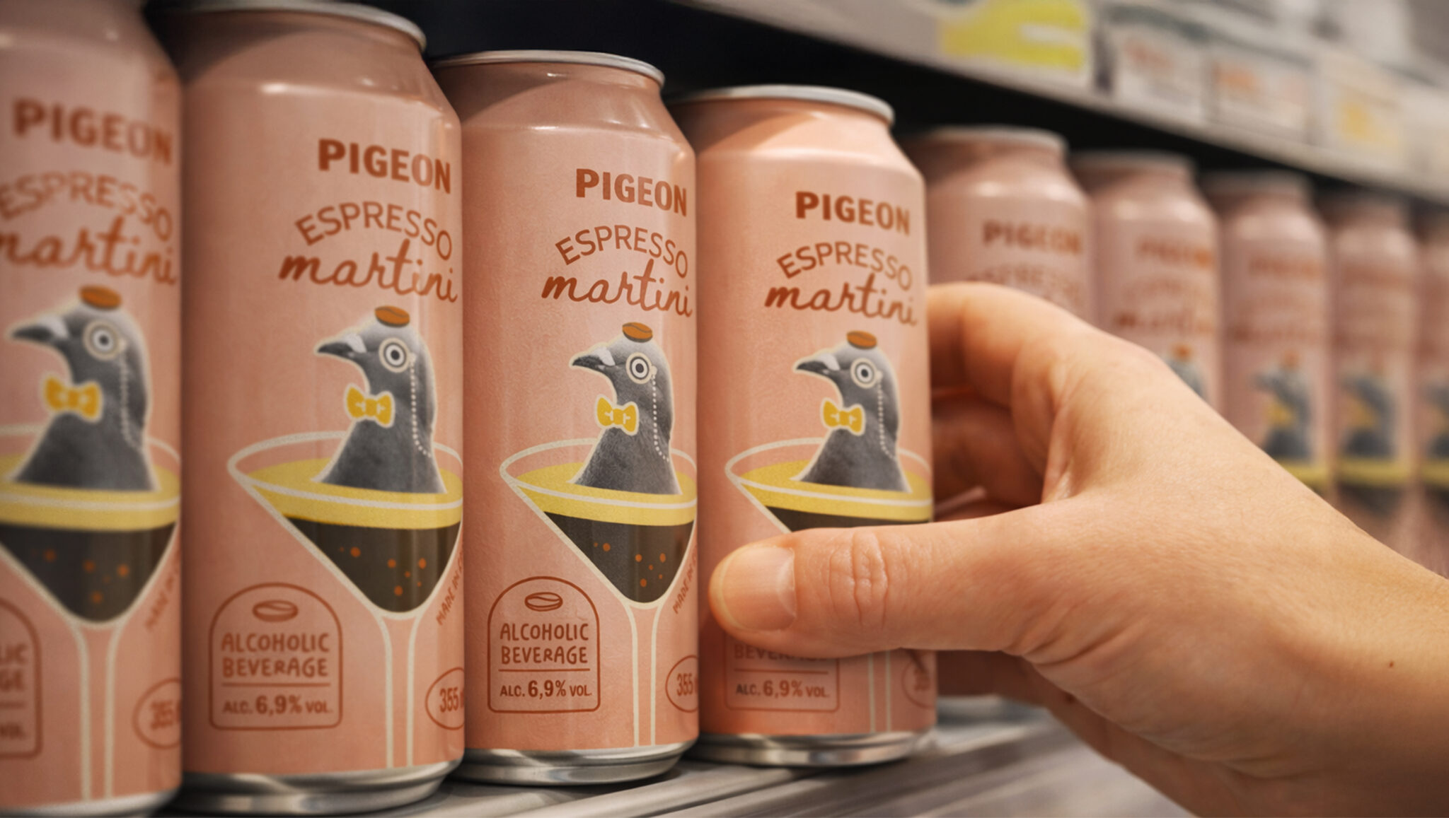 Une main saisit une canette de « Pigeon Espresso Martini » sur le rayon d'un magasin, au milieu d'autres canettes similaires ornées d'une illustration représentant un pigeon dans un verre à martini.