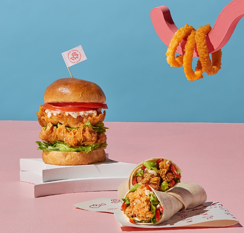 Un sandwich au poulet frit, garni de laitue et de tomate, est posé à côté d'un wrap au poulet découpé sur un support rose. À l'arrière-plan, une sculpture en forme de main tient des rondelles d'oignon sur un fond bleu.