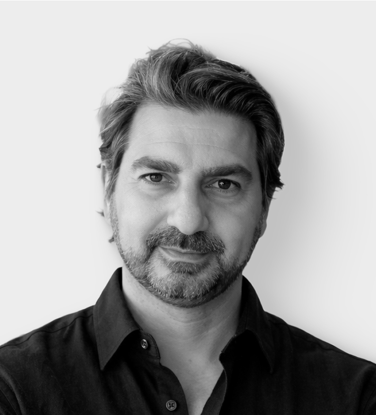 Un homme aux cheveux courts, foncés et ondulés, portant une barbe, vêtu d'une chemise sombre boutonnée, regarde directement la caméra avec un léger sourire, sur un fond clair et uni.