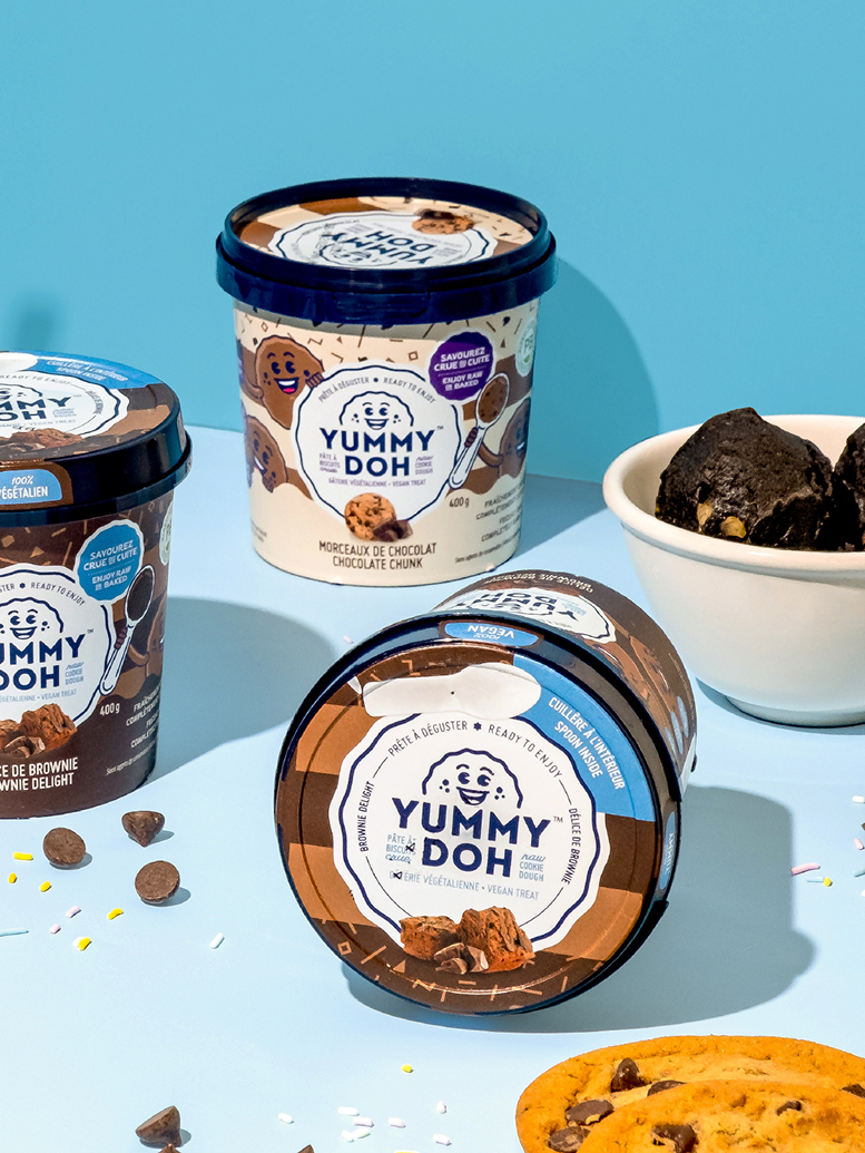Trois pots de pâte à biscuits comestible « Yummy Doh » sont disposés sur un support bleu et jaune, avec à côté des pépites de chocolat, un biscuit aux pépites de chocolat et un bol de pâte à biscuits. L'emballage est orné d'illustrations ludiques.