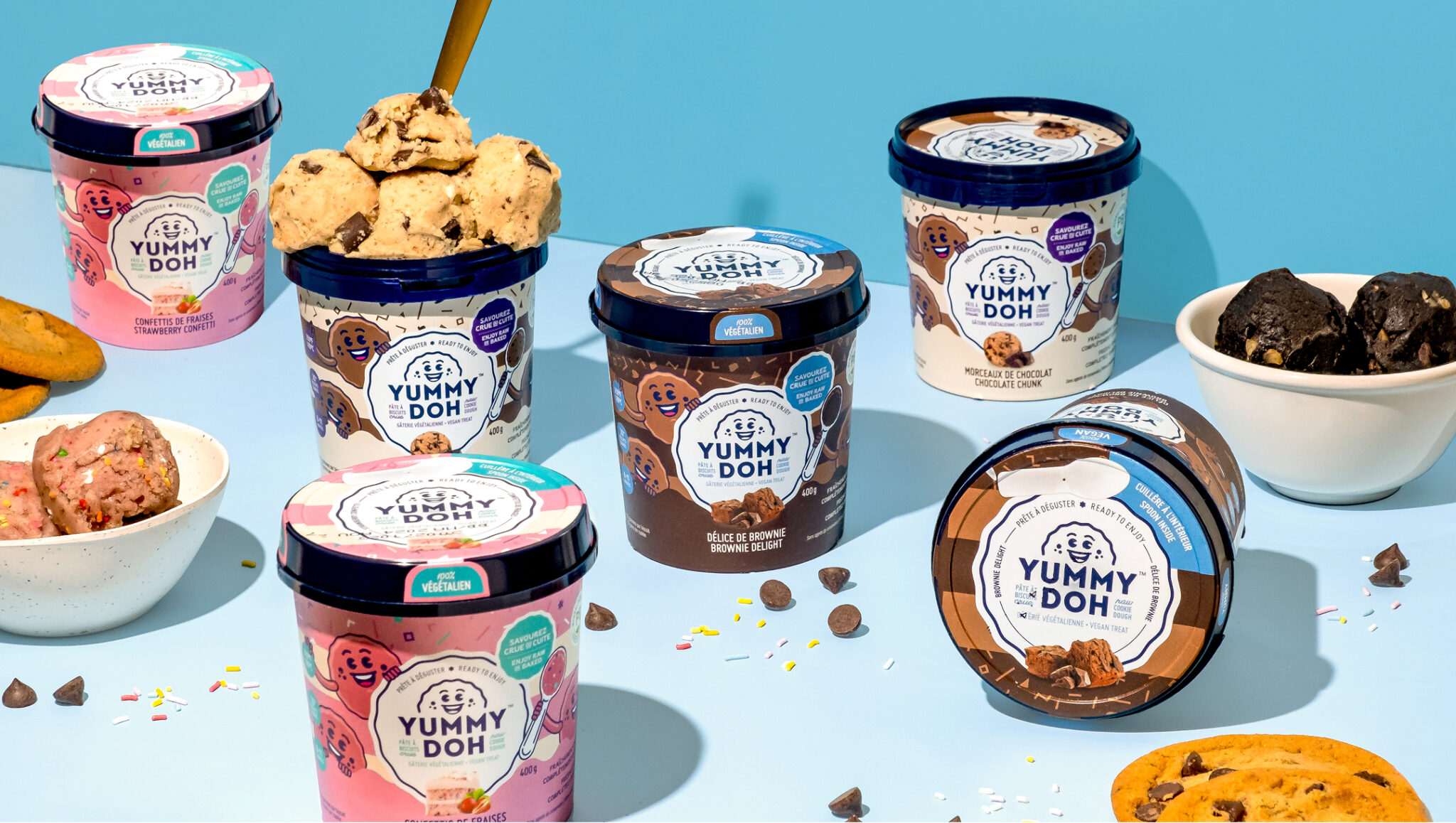 Plusieurs pots de pâte à biscuits comestible « Yummy Doh », de différentes saveurs, sont disposés sur un fond bleu, entourés de bols, de biscuits, de pépites de chocolat et de vermicelles en sucre éparpillés. Sur l'un des pots, des boules de pâte sont empilées.