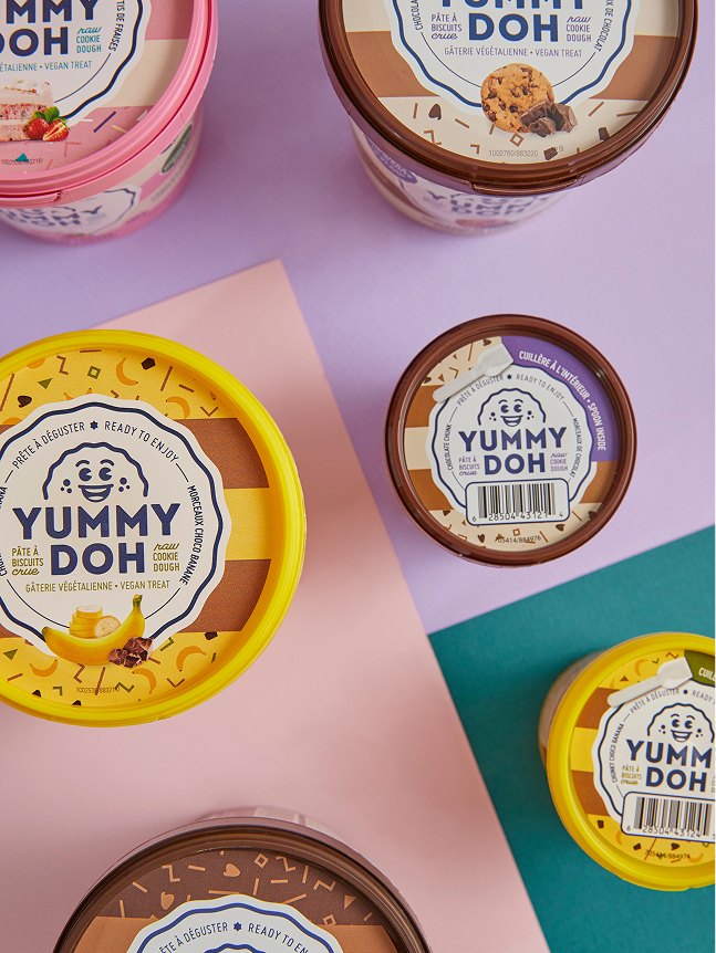Des pots assortis de pâte à biscuits végane « Yummy Doh » sont disposés sur un fond rose pastel et violet, avec des couvercles jaunes, roses et marron arborant le logo ludique de la marque ainsi que les informations sur le produit.